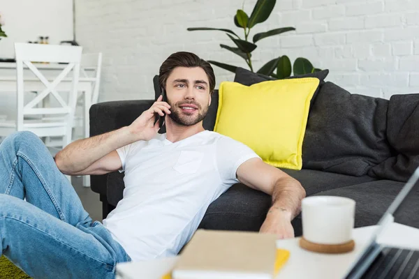 Smiling man talking on smartphone and sitting on floor near couch — Stock Photo