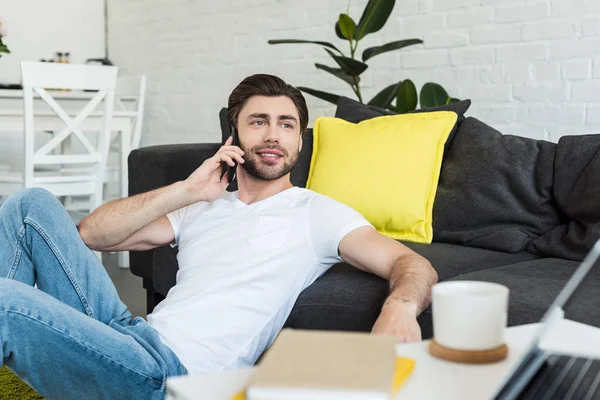 Hombre sonriente hablando en el teléfono inteligente y sentado en el suelo cerca del sofá - foto de stock