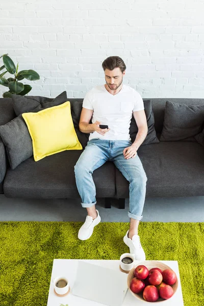 Aus der Vogelperspektive: junger Mann sitzt auf Sofa mit Smartphone in der Hand am Tisch mit Laptop, Äpfeln und Kaffeetassen zu Hause — Stockfoto