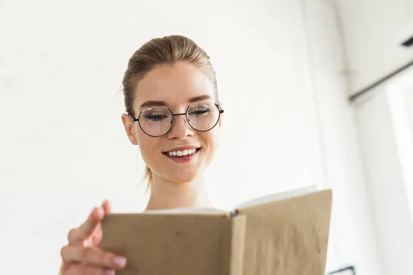 Tiefansicht einer jungen lächelnden Frau mit Brille beim Lesen eines Buches — Stockfoto