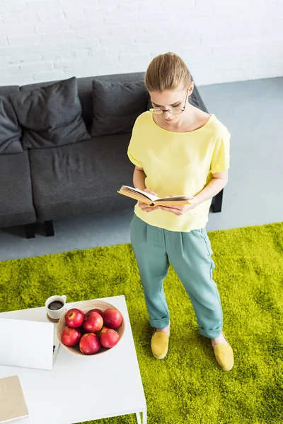 Visão de alto ângulo da jovem mulher em óculos livro de leitura perto da mesa com laptop, maçãs e xícara de café em casa — Fotografia de Stock