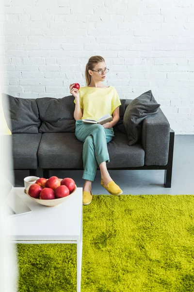 Junge Frau in Brille liest Buch und hält Apfel auf Sofa neben Tisch mit Kaffee, Äpfeln und Laptop zu Hause — Stockfoto