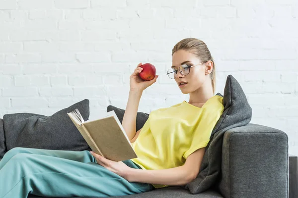 Jeune femme en lunettes tenant pomme et livre de lecture sur canapé à la maison — Photo de stock