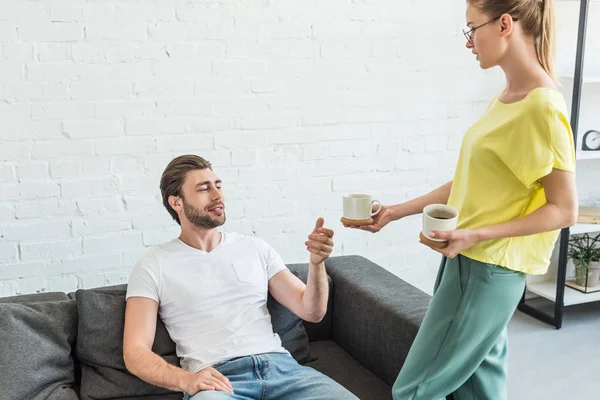 Vista lateral da jovem mulher em óculos dando xícara de café para namorado no sofá em casa — Fotografia de Stock