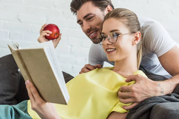 Молодий усміхнений чоловік обіймає дівчину, читаючи книгу з яблуком в руці на дивані — стокове фото