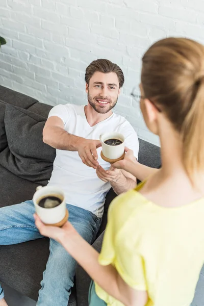 Vista posteriore della donna che dà tazza di caffè al fidanzato sorridente seduto sul divano a casa — Foto stock
