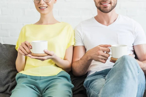 Cropped shot of smiling young couple holding coffee cups on sofa at home — Stock Photo