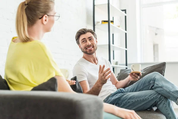 Joven hombre sonriente con taza de café gesticulando a mano y hablando con la novia en casa - foto de stock