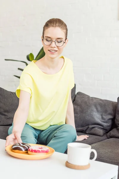 Lächelnde junge Frau mit Brille, die daheim Donuts vom Teller auf den Tisch mit Kaffeetasse holt — Stockfoto