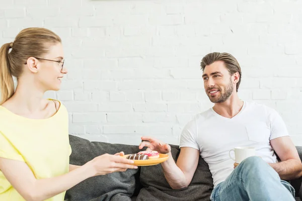 Smiling woman in eyeglasses offering donuts to boyfriend with coffee cup at home — Stock Photo