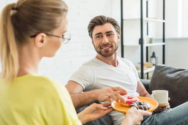 Mujer en gafas con plato de rosquillas mientras su novio toma una rosquilla y se sienta en el sofá en casa - foto de stock