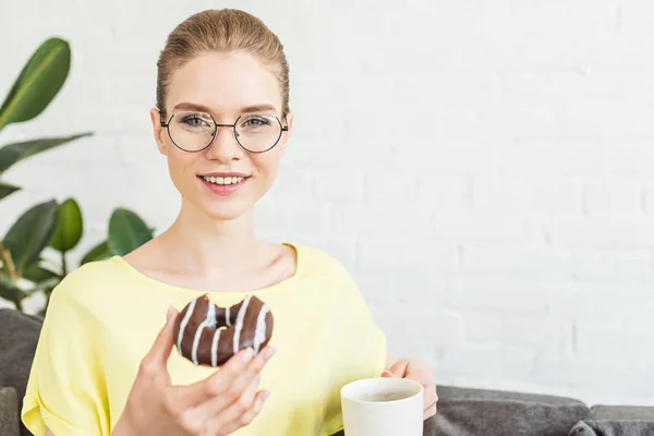 Sorridente giovane donna in occhiali bere caffè e mangiare ciambella a casa — Foto stock