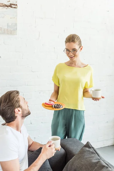 Lächelnder Mann im Gespräch mit Freundin mit Kaffeetasse und Teller mit Donuts zu Hause — Stockfoto