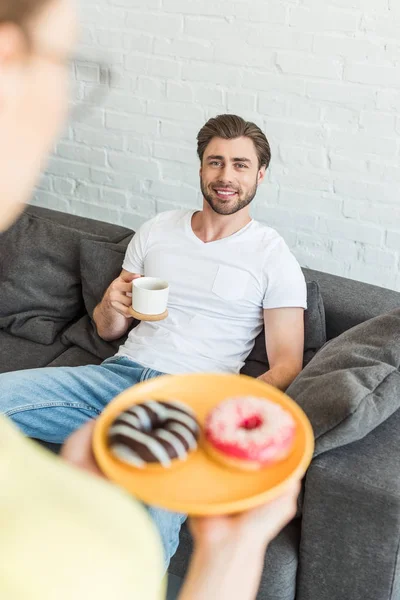 Schnappschuss einer Frau mit Donuts auf dem Teller und einem lächelnden Freund, der zu Hause auf dem Sofa mit Kaffeetasse sitzt — Stockfoto