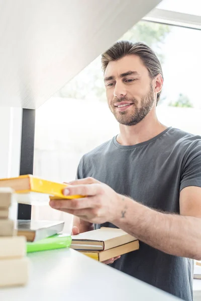 Giovane uomo sorridente mettere libri sugli scaffali a casa — Foto stock