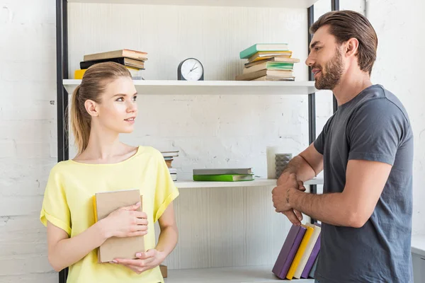 Young attractive woman holding book and talking to smiling boyfriend near shelves with books — Stock Photo