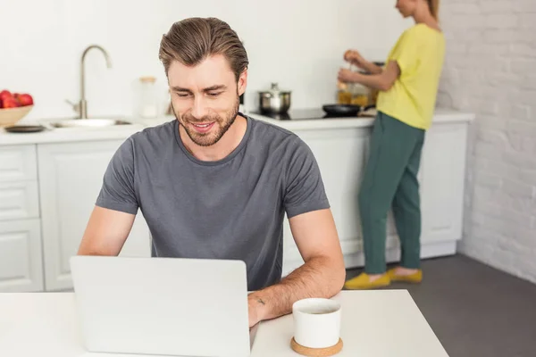 Sonriente joven trabajando en el ordenador portátil y su novia cocinando detrás en la cocina - foto de stock