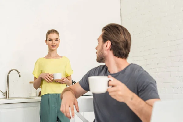 Joven con taza de café sentado en la silla y hablando con su novia en la cocina - foto de stock