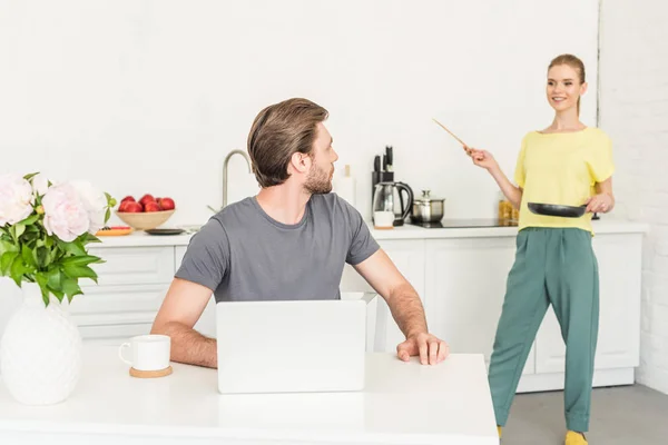 Молодой человек работает за столом с ноутбуком и его девушка стоит позади с сковородкой и лопаткой — стоковое фото