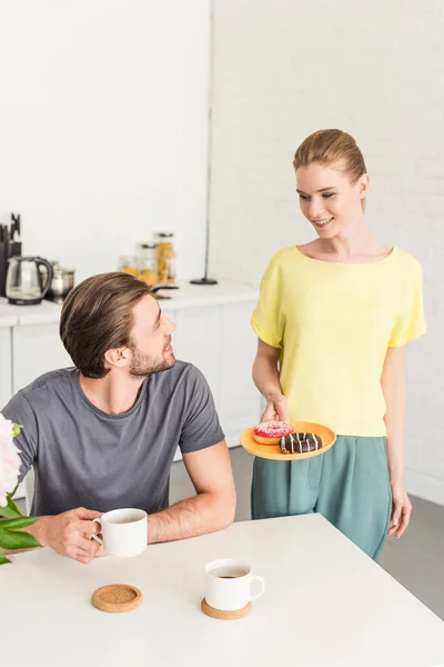 Smiling woman holding plate with donuts and talking to smiling boyfriend sitting at table — Stock Photo