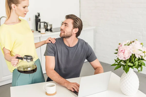 Atractiva mujer joven con café tocando hombro novio mientras él sentado en la mesa con el ordenador portátil - foto de stock