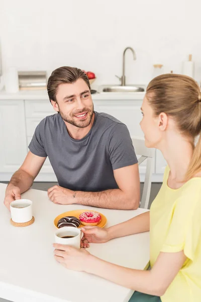 Joven sonriente pareja teniendo conversación en cocina mesa con rosquillas y tazas de café - foto de stock