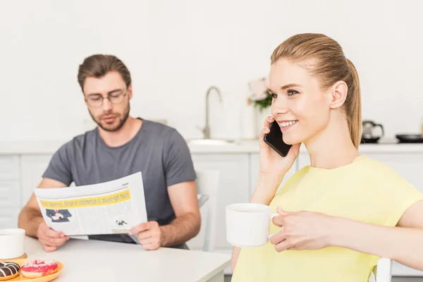 Sonriente mujer joven hablando en el teléfono inteligente y beber tazas de café mientras su novio leyendo el periódico en la cocina - foto de stock