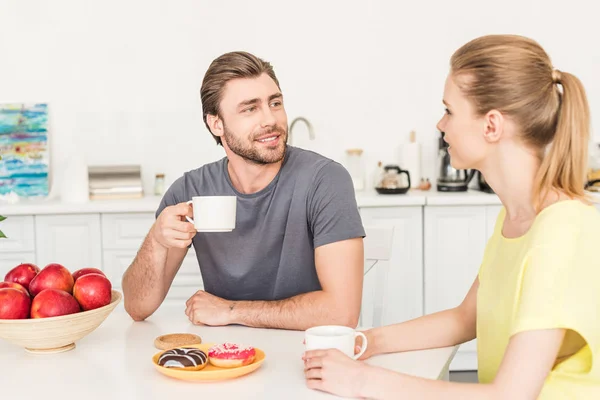 Молода пара розмовляє і снідає за столом з пончиками, чашками кави та яблуками — стокове фото