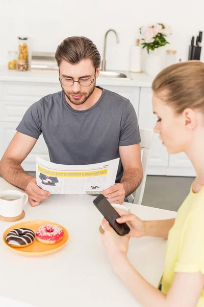 Vista lateral da mulher usando smartphone e namorado ler jornal à mesa com xícaras de café e donuts na cozinha — Fotografia de Stock