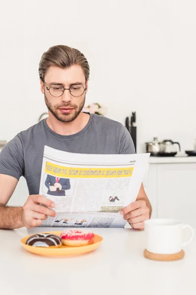 Man in eyeglasses reading newspaper at table with donuts and coffee cup — Stock Photo