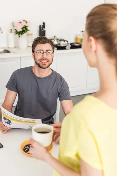 Молодой улыбающийся мужчина с газетой разговаривает с подругой с чашкой кофе на кухне — стоковое фото
