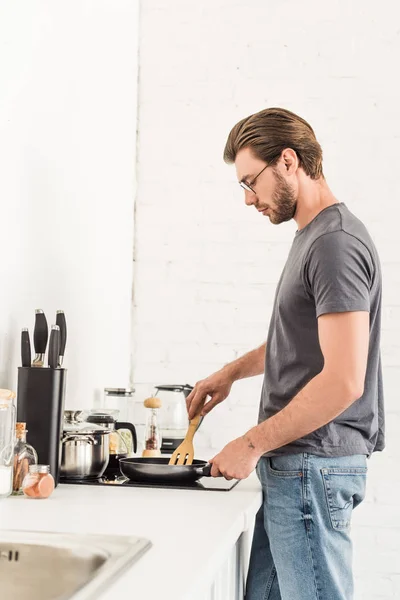 Vue latérale du jeune homme cuisinier sur cuisinière avec poêle et spatule à la cuisine — Photo de stock