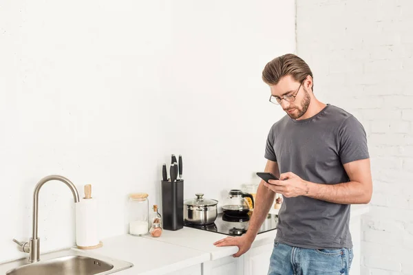 Visão frontal do jovem em óculos verificando smartphone na cozinha — Fotografia de Stock