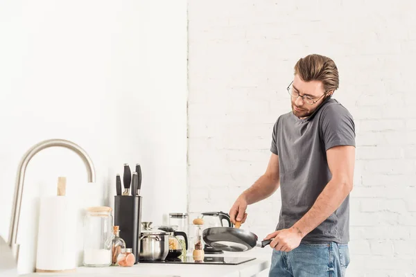 Young man talking on smartphone and cooking with frying pan and spatula at kitchen — Stock Photo