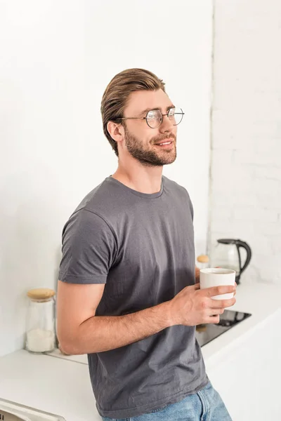 Smiling young man in eyeglasses drinking coffee at kitchen — Stock Photo