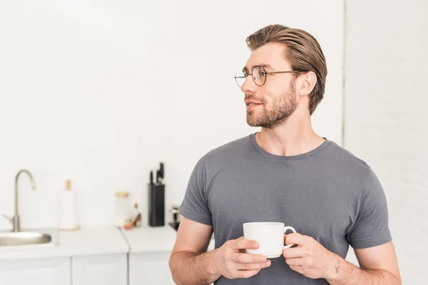 Side view of smiling young man in eyeglasses drinking coffee at kitchen — Stock Photo