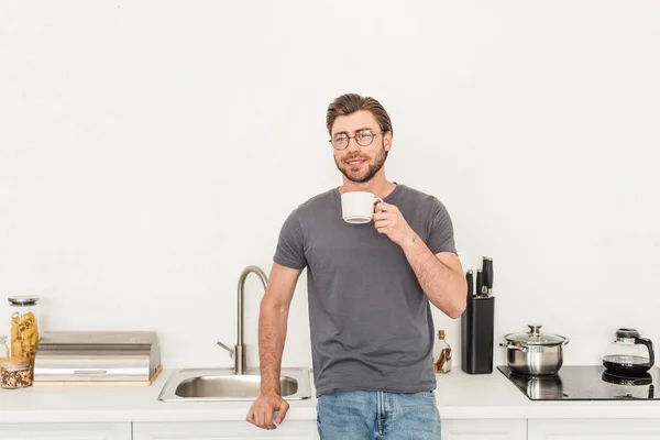 Front view of smiling young man in eyeglasses drinking coffee at kitchen — Stock Photo