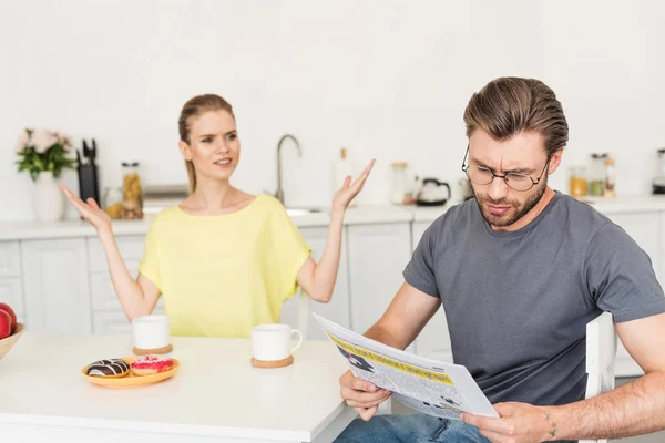 Young woman with wide arms looking at boyfriend while he reading newspaper at table with breakfast — Stock Photo