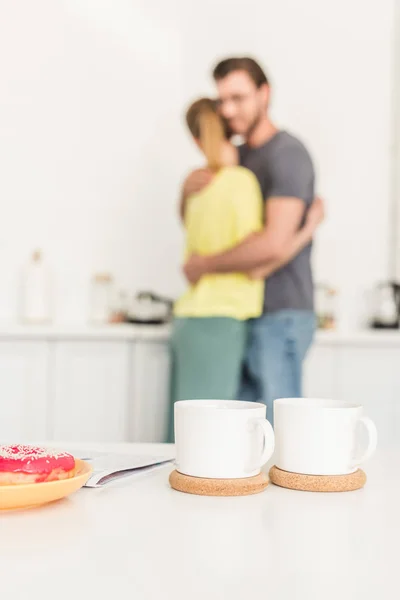 Close-up tiro de duas xícaras de café à mesa com donuts e casal abraçando atrás na cozinha — Fotografia de Stock