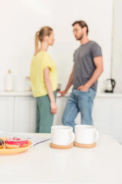 Close up view of two coffee cups at table with donuts and couple standing behind at kitchen — Stock Photo