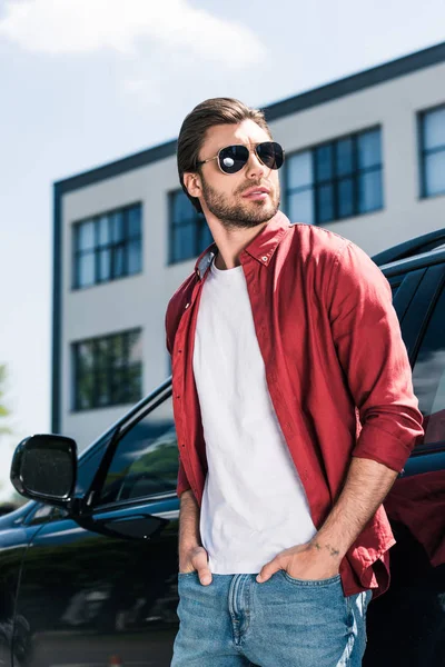 Élégant modèle masculin en lunettes de soleil avec les mains dans les poches debout près de voiture noire — Photo de stock