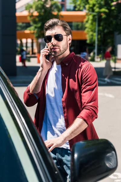 Stylish man in sunglasses talking on smartphone and opening door of own car at urban street — Stock Photo