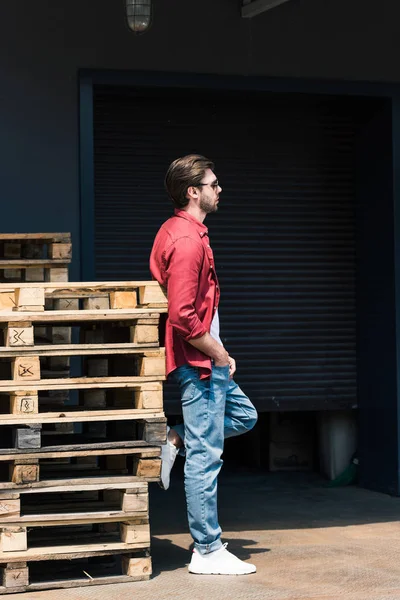 Vista laterale di giovane uomo elegante in occhiali da sole in piedi vicino pallet di legno — Foto stock