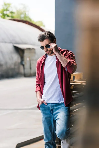 Front view of young stylish man standing near wooden pallets and adjusting sunglasses — Stock Photo