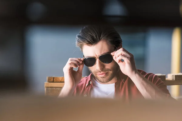 Stilvoller junger Mann steht neben Holzpaletten und justiert Sonnenbrille — Stockfoto