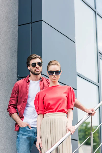Front view of stylish young couple of models in sunglasses standing on staircases — Stock Photo