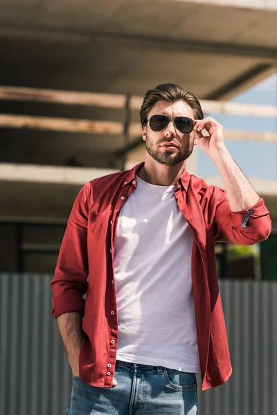 Front view of young stylish man adjusting sunglasses in front of construction building — Stock Photo