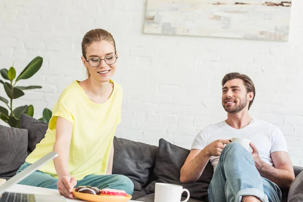 Joven pareja sonriente desayunando con café y rosquillas en el sofá en casa - foto de stock