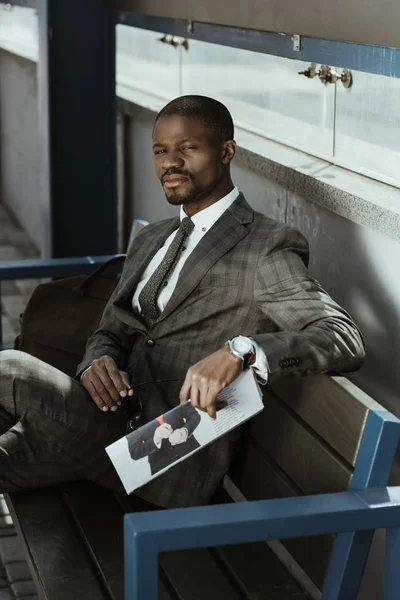 Jovem empresário afro-americano sentado no banco com jornal — Fotografia de Stock