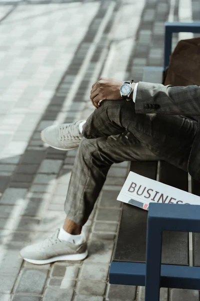 Cropped view of African american businessman wearing suit sitting on bench with newspaper — Stock Photo